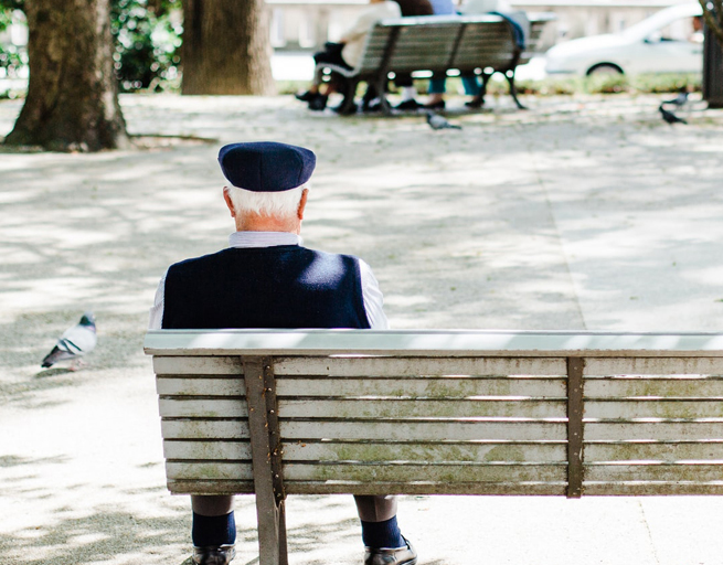 Photo of elderly gent sitting on a park bench in blue vest and cap